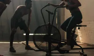Träning med assault bike intervaller. En kvinna cyklar intensivt medan en man vilar, utmattad efter passet.
