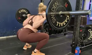 Kvinna gör squats med skivstång i ett gym. Hon har på sig bruna träningskläder och orangea skor.
