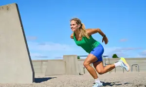 Kvinna i grönt linne och blå shorts utför ett intensivt hiit pass på en sandig yta, med betongkonstruktioner i bakgrunden.