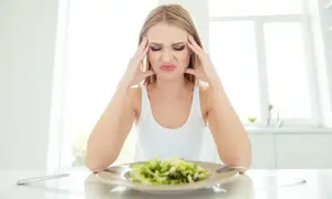 Kvinna med huvudvärk och grimas framför en sallad, troliga symptom på kaloriunderskott.