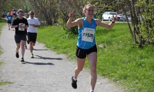 Glad löpare med nummer 11526, Elin, vinkar glatt under ett maraton. Hon har precis passerat andra löpare på grusvägen.