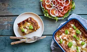 En bit nyttig lasagne på en tallrik bredvid en hel lasagneform och en fräsch tomatsallad med rödlök.