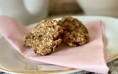 Tre hembakade, kalorisnåla kakor med havre och chokladbitar ligger på en ljusrosa servett på en vit tallrik.
