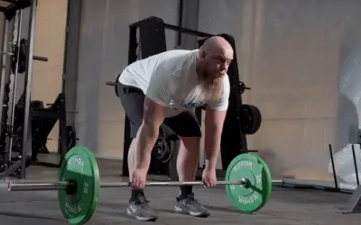 Man med skägg utför raka marklyft med en skivstång med gröna vikter.