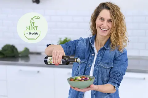 Glad kvinna häller olivolja över en fräsch sallad. En logotyp med texten "ÄT SUND - för alltid" syns i bakgrunden, vilket antyder en livsstil med fet mat och hälsosamma val.