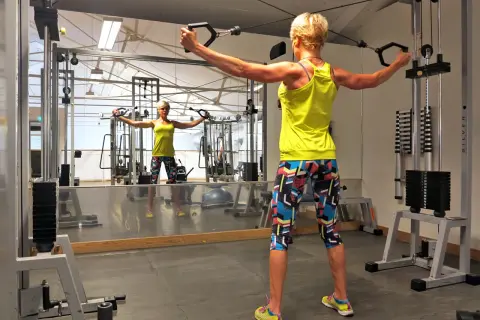 Kvinna tränar baksida axlar med kabelmaskin i gym. Spegeln visar hennes reflektion.