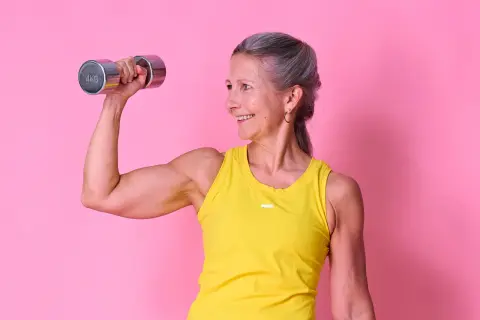 Glad kvinna med grått hår lyfter en 4 kg hantel, visar upp sina muskler. Hon inspirerar till frågan: hur mycket muskler kan man bygga på ett år?