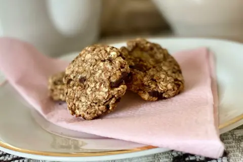Tre hembakade, kalorisnåla kakor med havre och chokladbitar ligger på en ljusrosa servett på en vit tallrik.