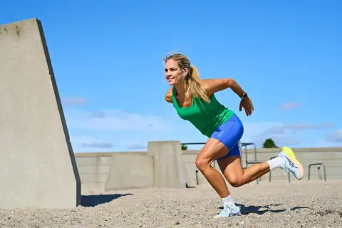 Kvinna i grönt linne och blå shorts utför ett intensivt hiit pass på en sandig yta, med betongkonstruktioner i bakgrunden.