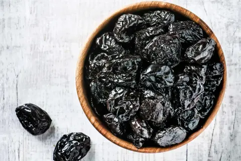 En skål med torkade plommon, som är nyttigt och gott. Några plommon ligger bredvid på en vit bakgrund.