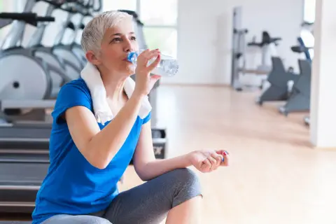 En kvinna med grått hår dricker ljummet vatten ur en flaska efter träningen på gymmet.