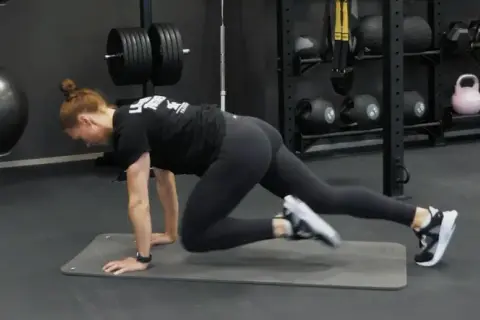 Kvinna gör mountain climbers på en träningsmatta i ett gym. Hon verkar ha dålig kondition, men kämpar på.