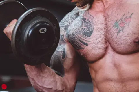 Muskulös man med tatueringar lyfter en 30 kg hantel. En del av resan för att gå ner i vikt och bygga muskler.