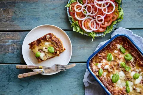 En bit nyttig lasagne på en tallrik bredvid en hel lasagneform och en fräsch tomatsallad med rödlök.