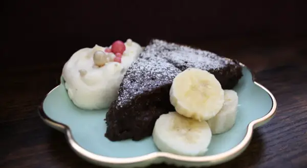 En bit nyttig kladdkaka med vispgrädde, bananskivor och strössel på en ljusblå tallrik.
