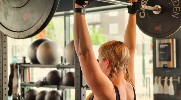 En kvinna lyfter en skivstång med vikter. Hon har flätor och träningshandskar. Frågan "hur blir man stark" besvaras med dedikation och hårt arbete på gymmet.