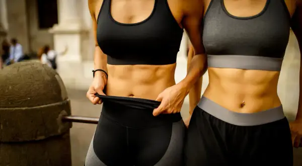 Två kvinnor visar upp sina vältränade magar. Frågan "hur får man six pack" besvaras med träning och dedikation.