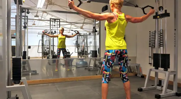 Kvinna tränar baksida axlar med kabelmaskin i gym. Spegeln visar hennes reflektion.