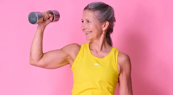 Glad kvinna med grått hår lyfter en 4 kg hantel, visar upp sina muskler. Hon inspirerar till frågan: hur mycket muskler kan man bygga på ett år?
