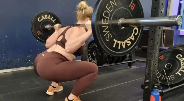 Kvinna gör squats med skivstång i ett gym. Hon har på sig bruna träningskläder och orangea skor.