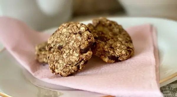 Tre hembakade, kalorisnåla kakor med havre och chokladbitar ligger på en ljusrosa servett på en vit tallrik.