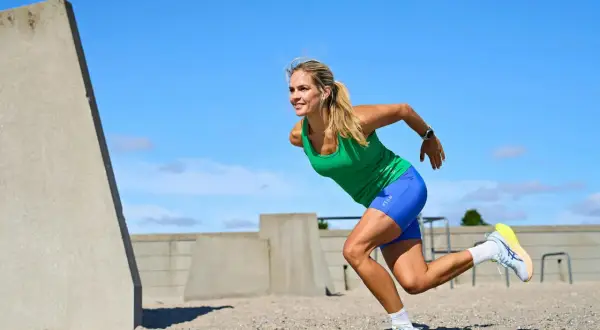 Kvinna i grönt linne och blå shorts utför ett intensivt hiit pass på en sandig yta, med betongkonstruktioner i bakgrunden.