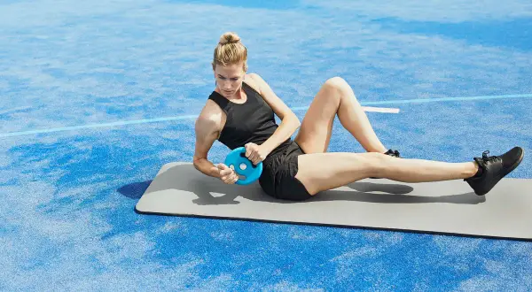 Kvinna gör sneda sit-ups med en blå viktplatta på en matta utomhus.