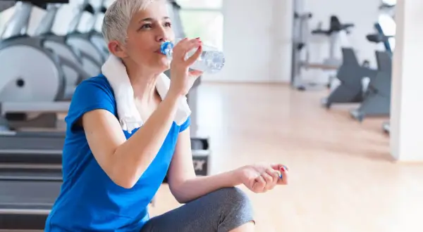 En kvinna med grått hår dricker ljummet vatten ur en flaska efter träningen på gymmet.