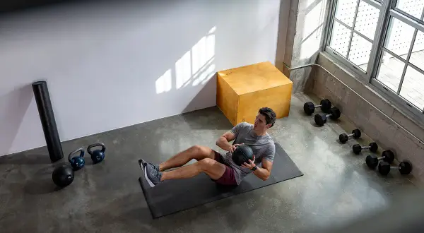 Man gör ett effektivt hiit pass hemma med en medicinboll. Träningsutrustning som kettlebells och hantlar syns i bakgrunden.
