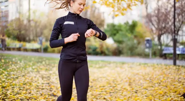 Kvinna springer i parken bland höstlöv och kollar sin klocka, hoppas på en bra tid på 5 km.