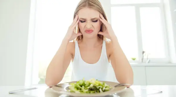 Kvinna med huvudvärk och grimas framför en sallad, troliga symptom på kaloriunderskott.
