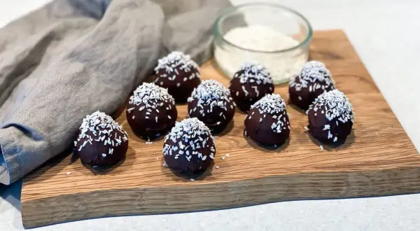 Mörka, chokladöverdragna protein chokladbollar toppade med kokosflingor ligger på en träskärbräda.