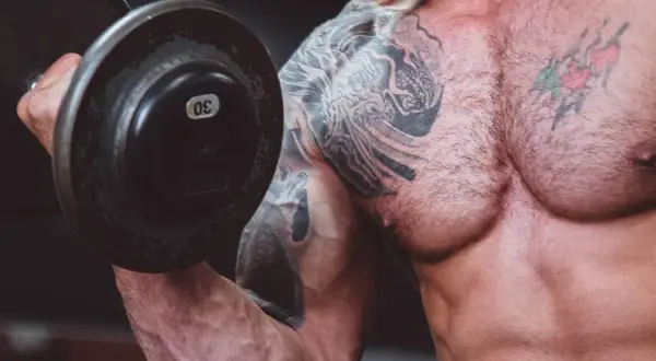 Muskulös man med tatueringar lyfter en 30 kg hantel. En del av resan för att gå ner i vikt och bygga muskler.