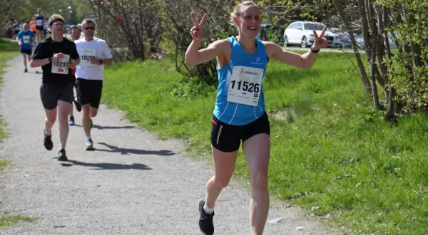 Glad löpare med nummer 11526, Elin, vinkar glatt under ett maraton. Hon har precis passerat andra löpare på grusvägen.