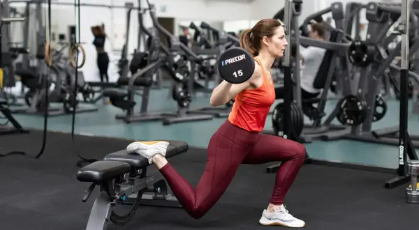 Kvinna utför en romanian split squat med skivstång på axlarna i ett gym.