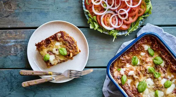 En bit nyttig lasagne på en tallrik bredvid en hel lasagneform och en fräsch tomatsallad med rödlök.