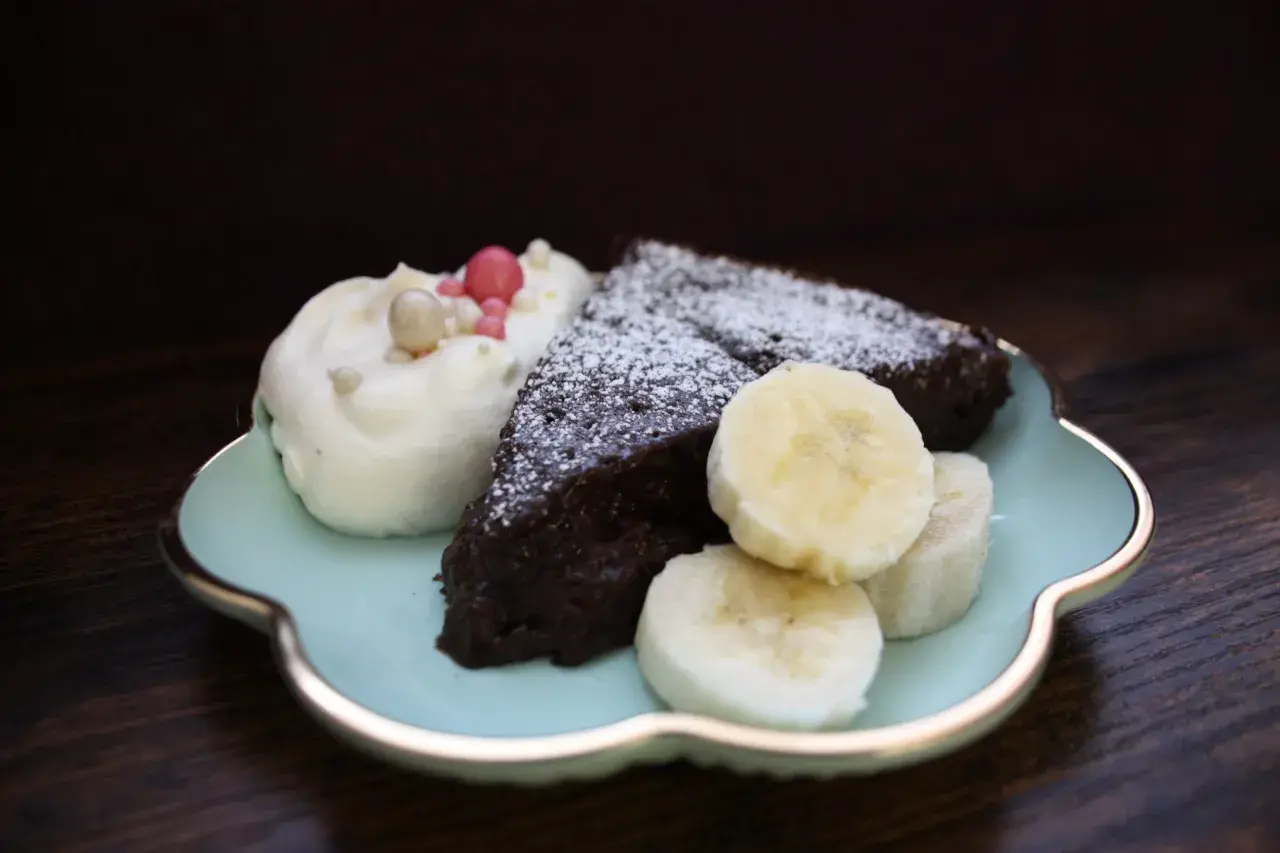En bit nyttig kladdkaka med vispgrädde, bananskivor och strössel på en ljusblå tallrik.