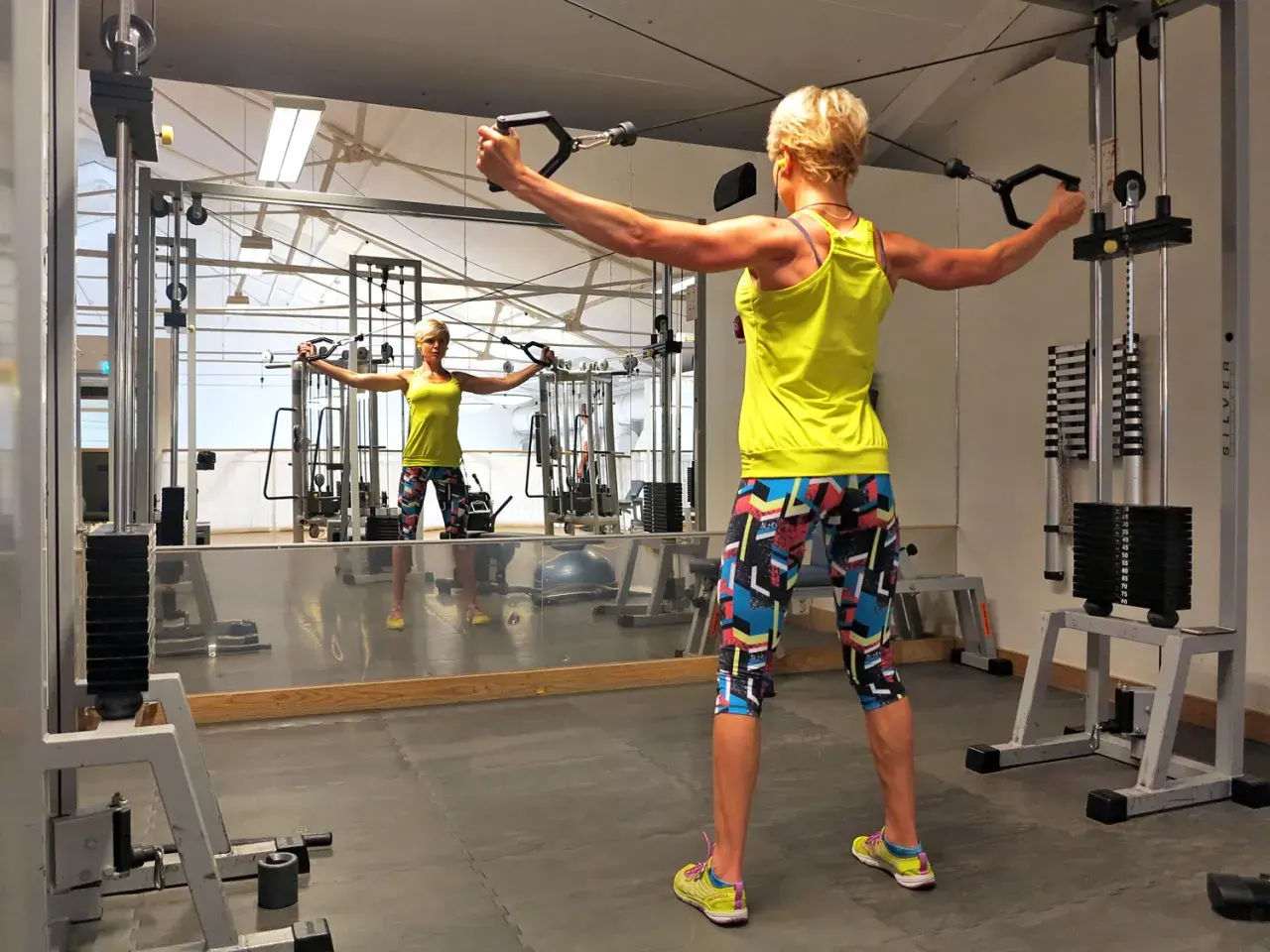 Kvinna tränar baksida axlar med kabelmaskin i gym. Spegeln visar hennes reflektion.