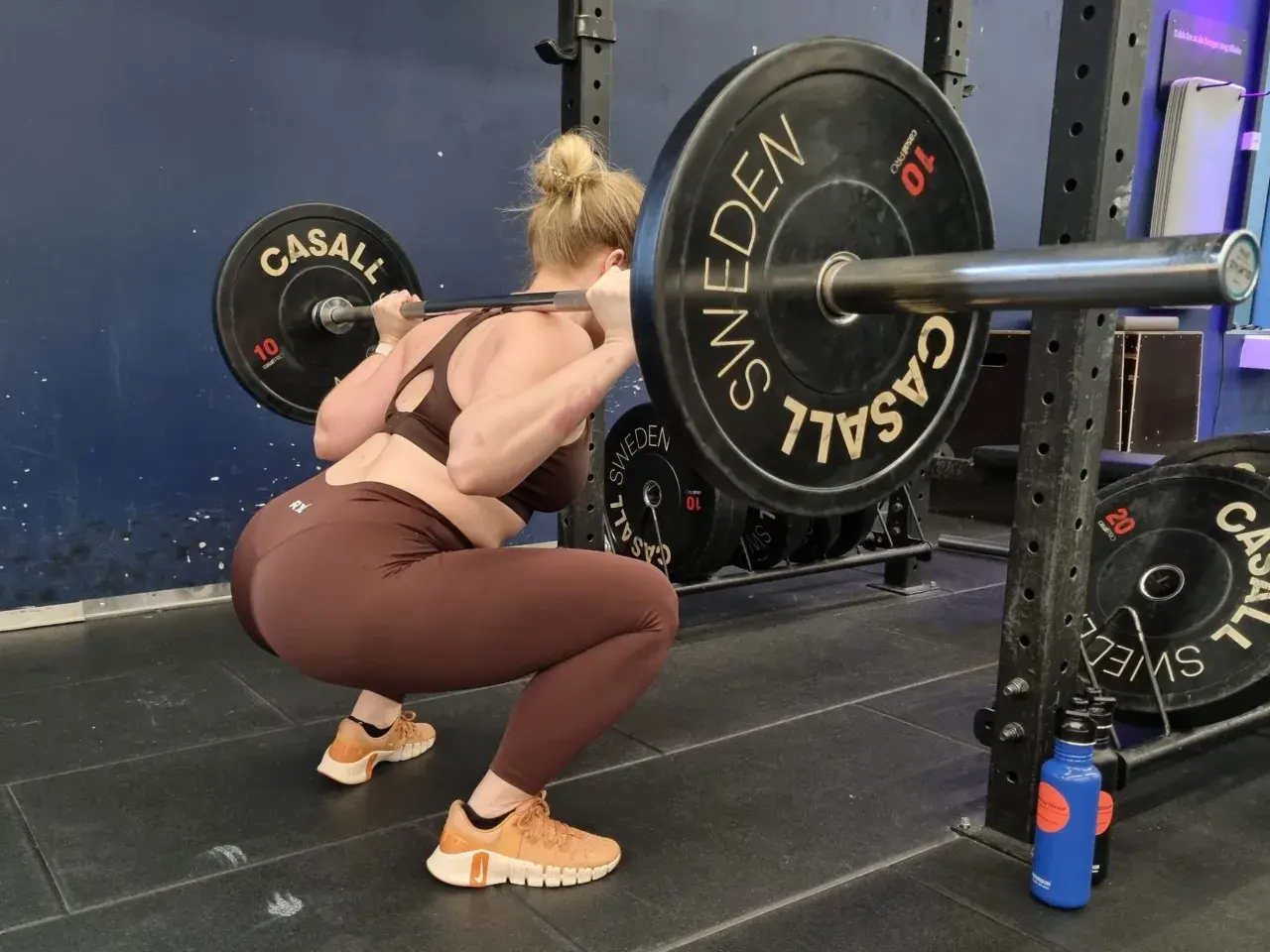 Kvinna gör squats med skivstång i ett gym. Hon har på sig bruna träningskläder och orangea skor.