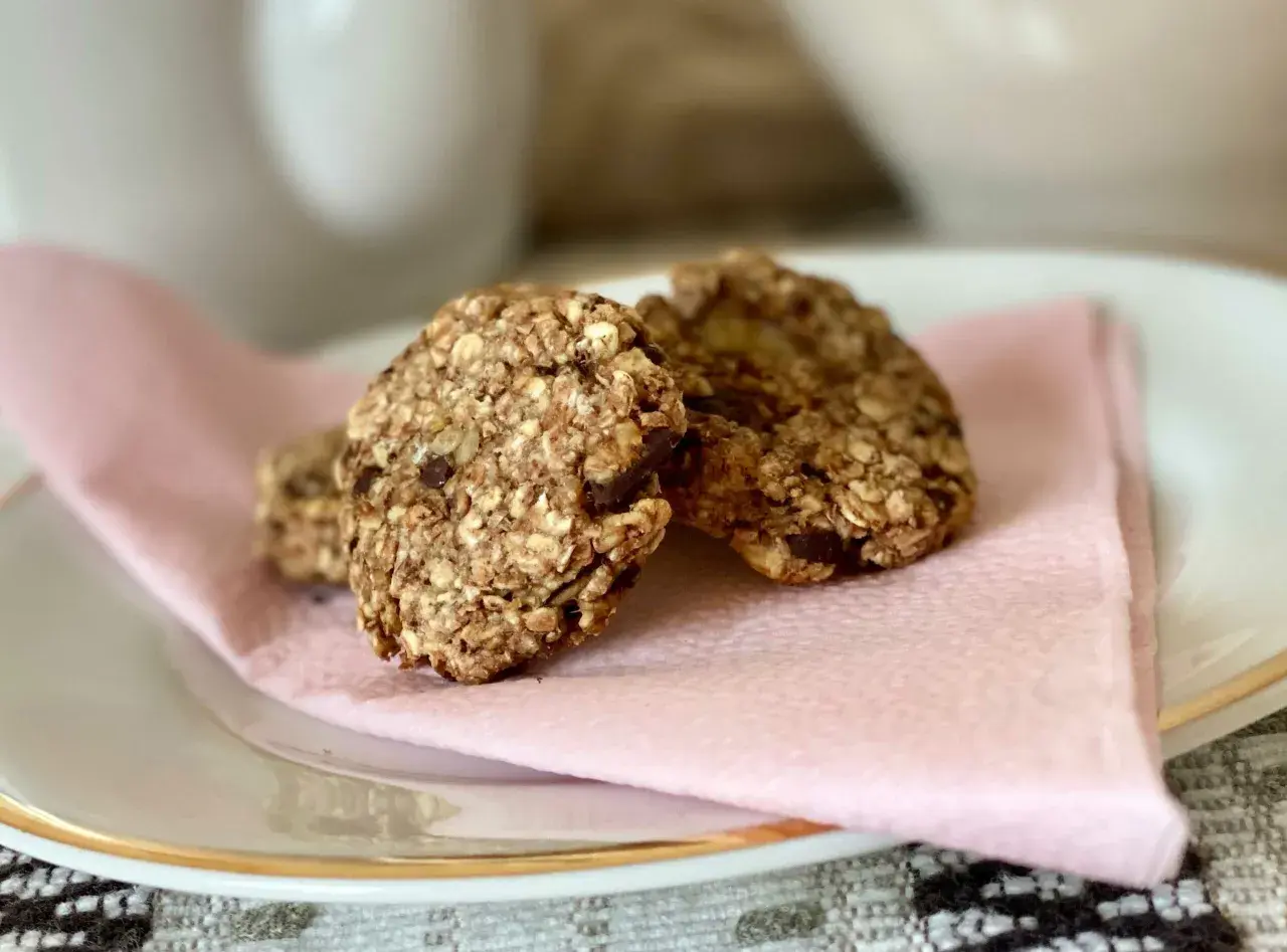 Tre hembakade, kalorisnåla kakor med havre och chokladbitar ligger på en ljusrosa servett på en vit tallrik.