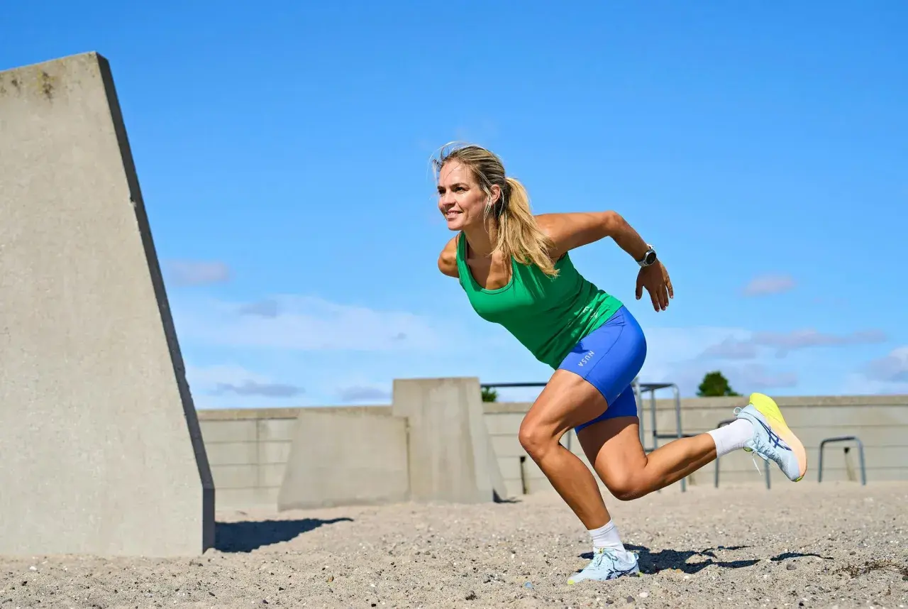 Kvinna i grönt linne och blå shorts utför ett intensivt hiit pass på en sandig yta, med betongkonstruktioner i bakgrunden.
