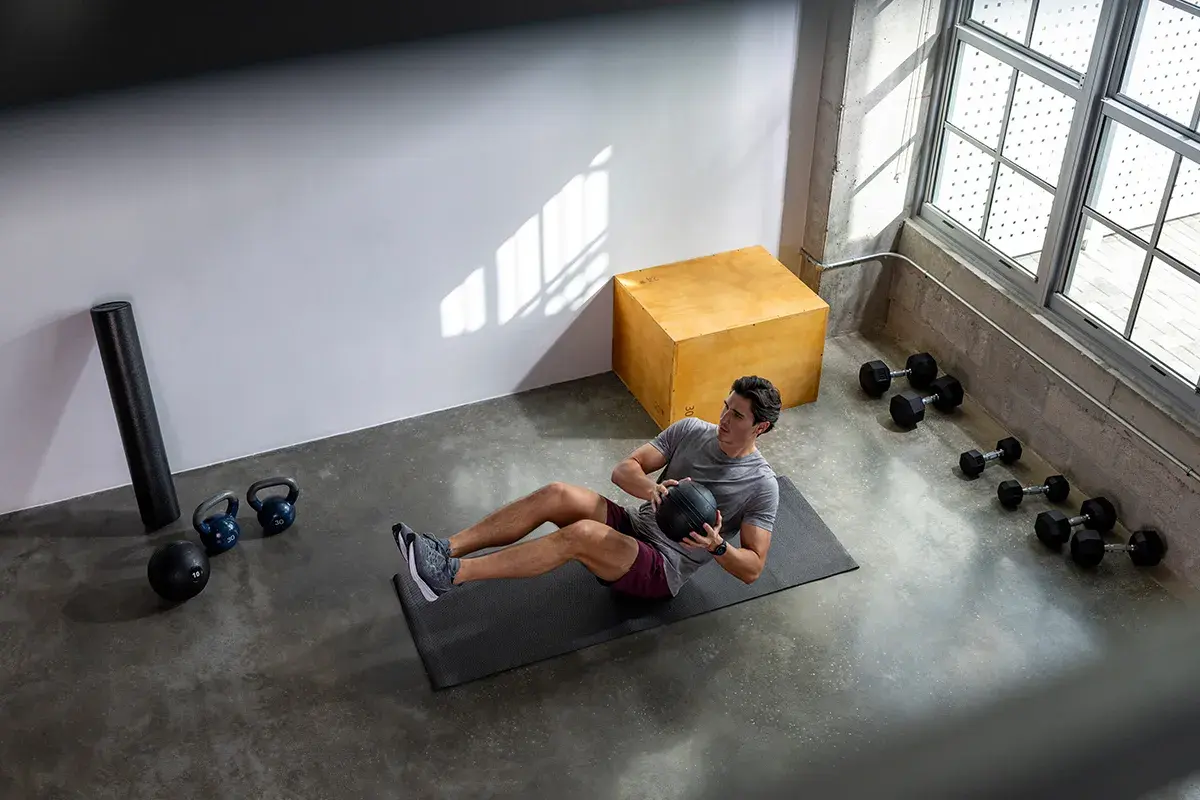 Man gör ett effektivt hiit pass hemma med en medicinboll. Träningsutrustning som kettlebells och hantlar syns i bakgrunden.