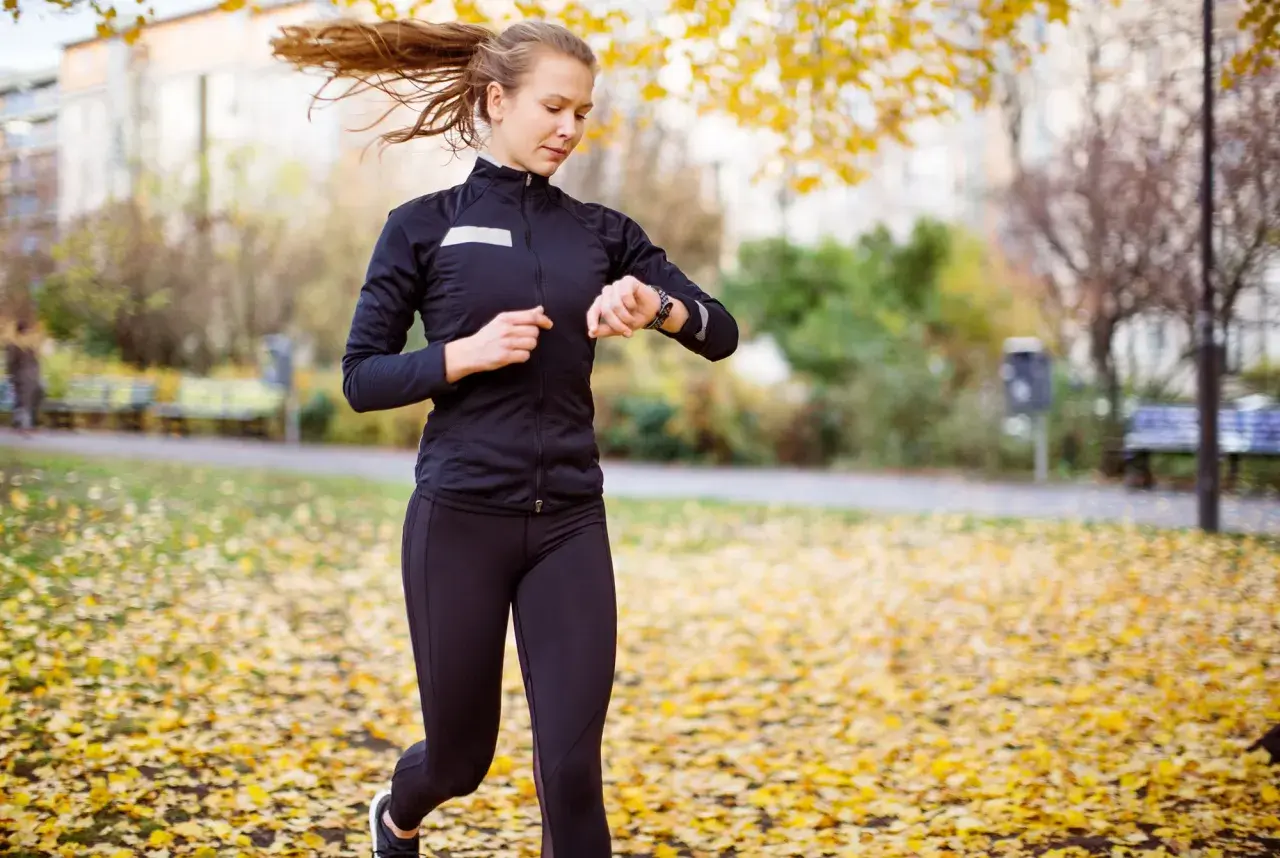Kvinna springer i parken bland höstlöv och kollar sin klocka, hoppas på en bra tid på 5 km.