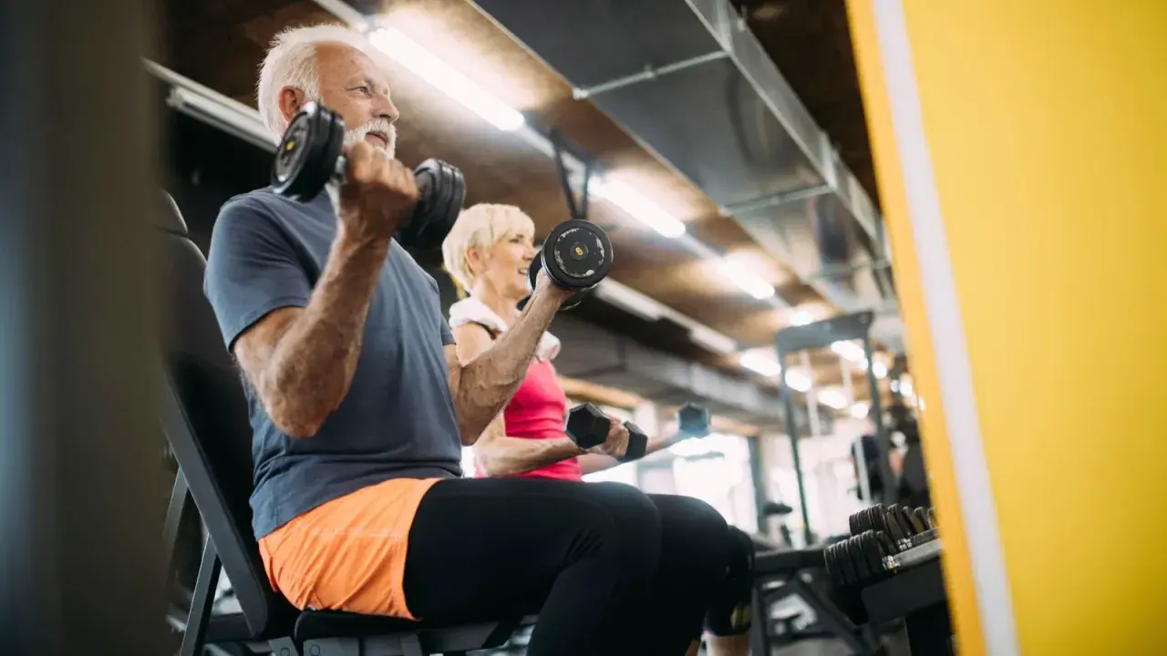 Äldre par tränar med hantlar på gym. De visar att sarkopeni betyder minskad muskelmassa, men att träning kan motverka detta.