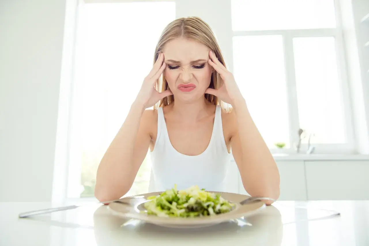 Kvinna med huvudvärk och grimas framför en sallad, troliga symptom på kaloriunderskott.