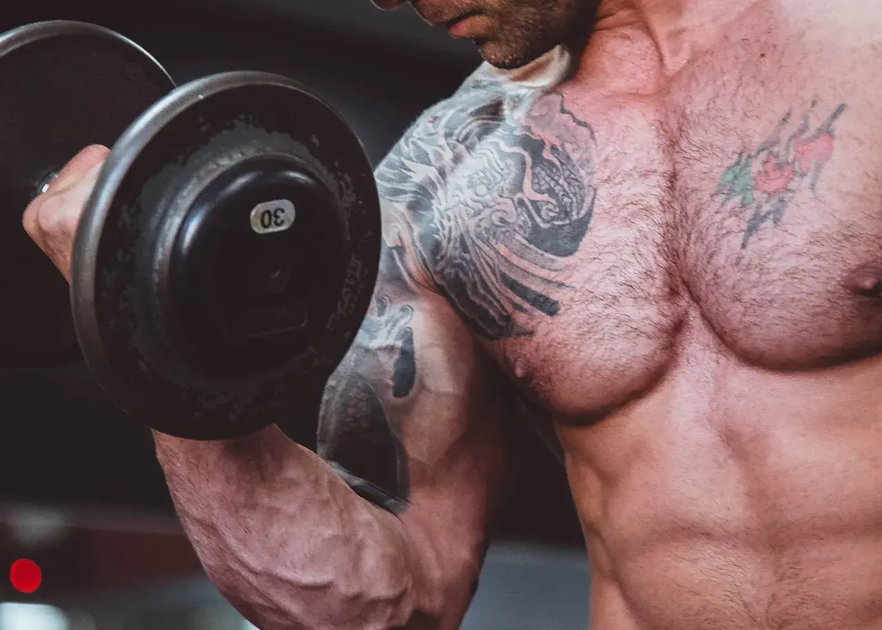 Muskulös man med tatueringar lyfter en 30 kg hantel. En del av resan för att gå ner i vikt och bygga muskler.