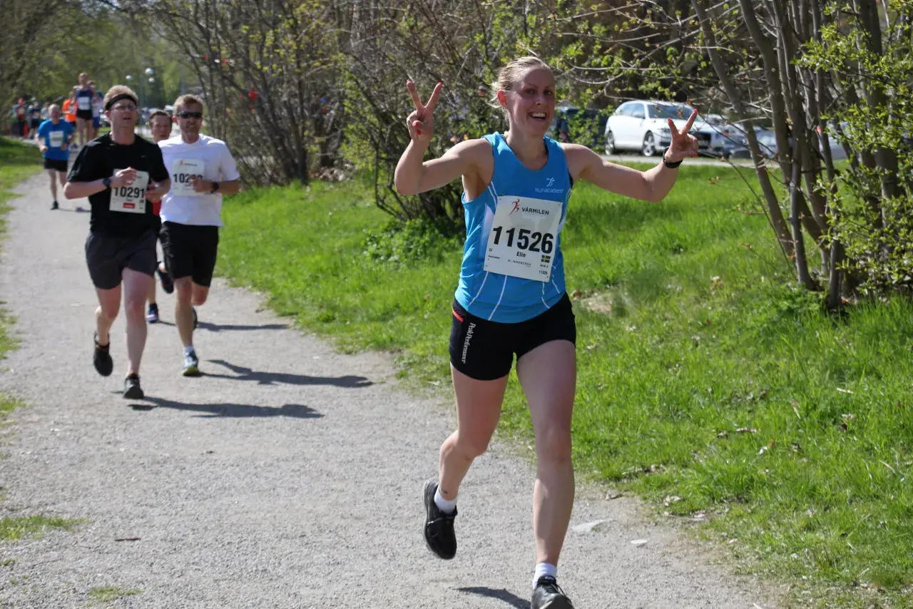 Glad löpare med nummer 11526, Elin, vinkar glatt under ett maraton. Hon har precis passerat andra löpare på grusvägen.