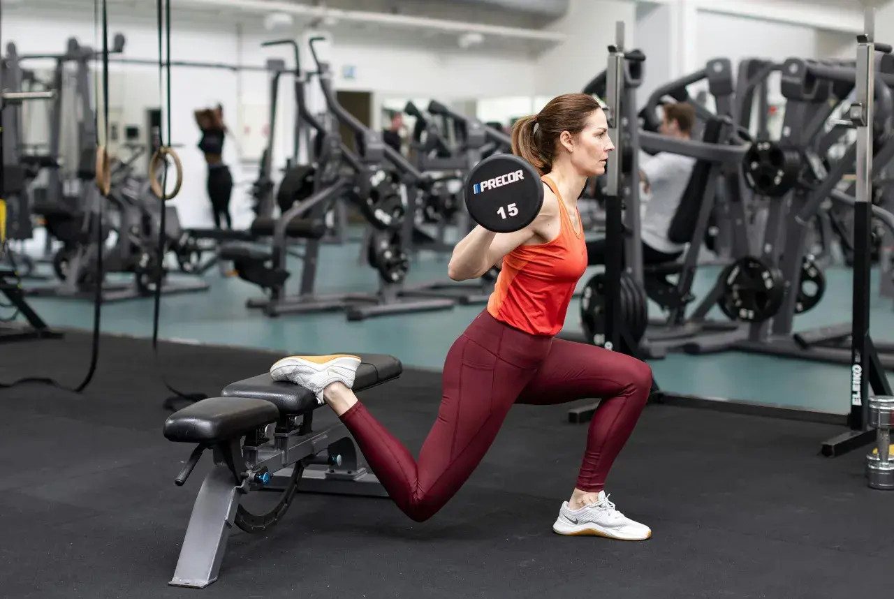 Kvinna utför en romanian split squat med skivstång på axlarna i ett gym.