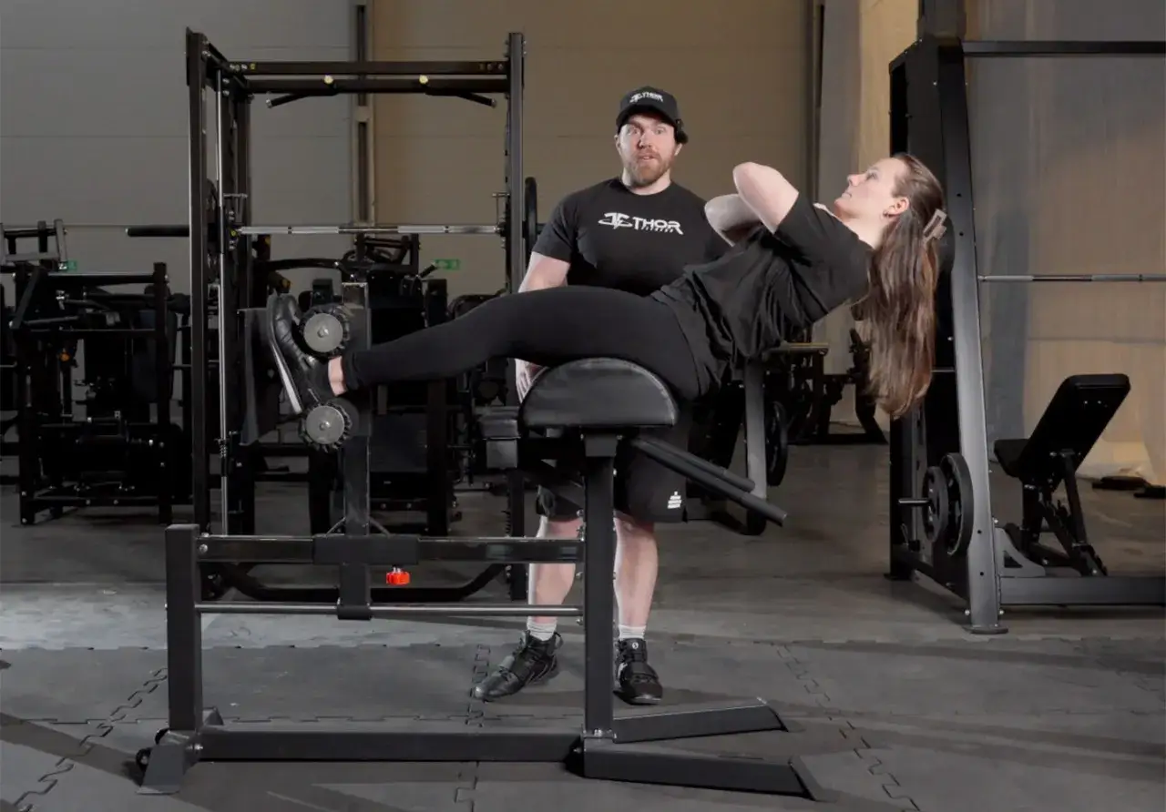 Man och kvinna tränar på gym. Kvinnan gör situps på en bänk, med fötterna fixerade i en maskin som liknar frog pumps.
