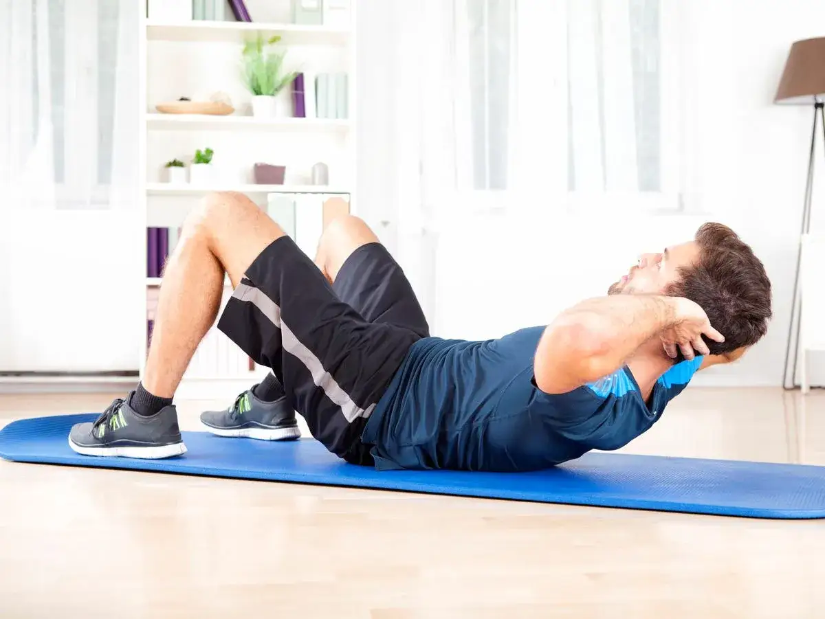 Man gör sit-ups på en blå träningsmatta i ett ljust rum med bokhyllor i bakgrunden.